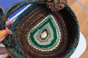 A top down view a small brown coiled pine needle basket with green binding. A large green stone makes up the decorative center and a small pinecone is attached to the rim near the handle.