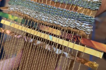 A small loom warped with shiny golden thread. Some fabric is woven with regular grey yarn, and strips of shiny copper are woven in periodically.